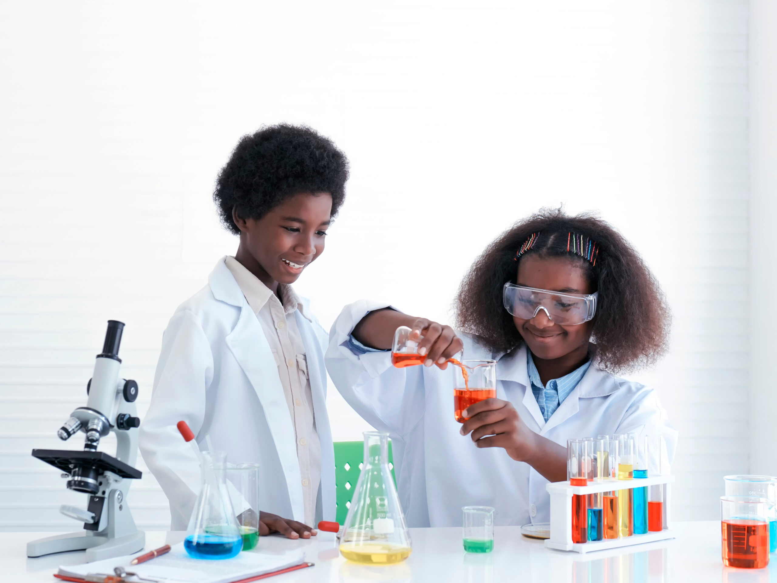 Two children pour brightly coloured liquids into beakers and flasks while wearing lab coats.